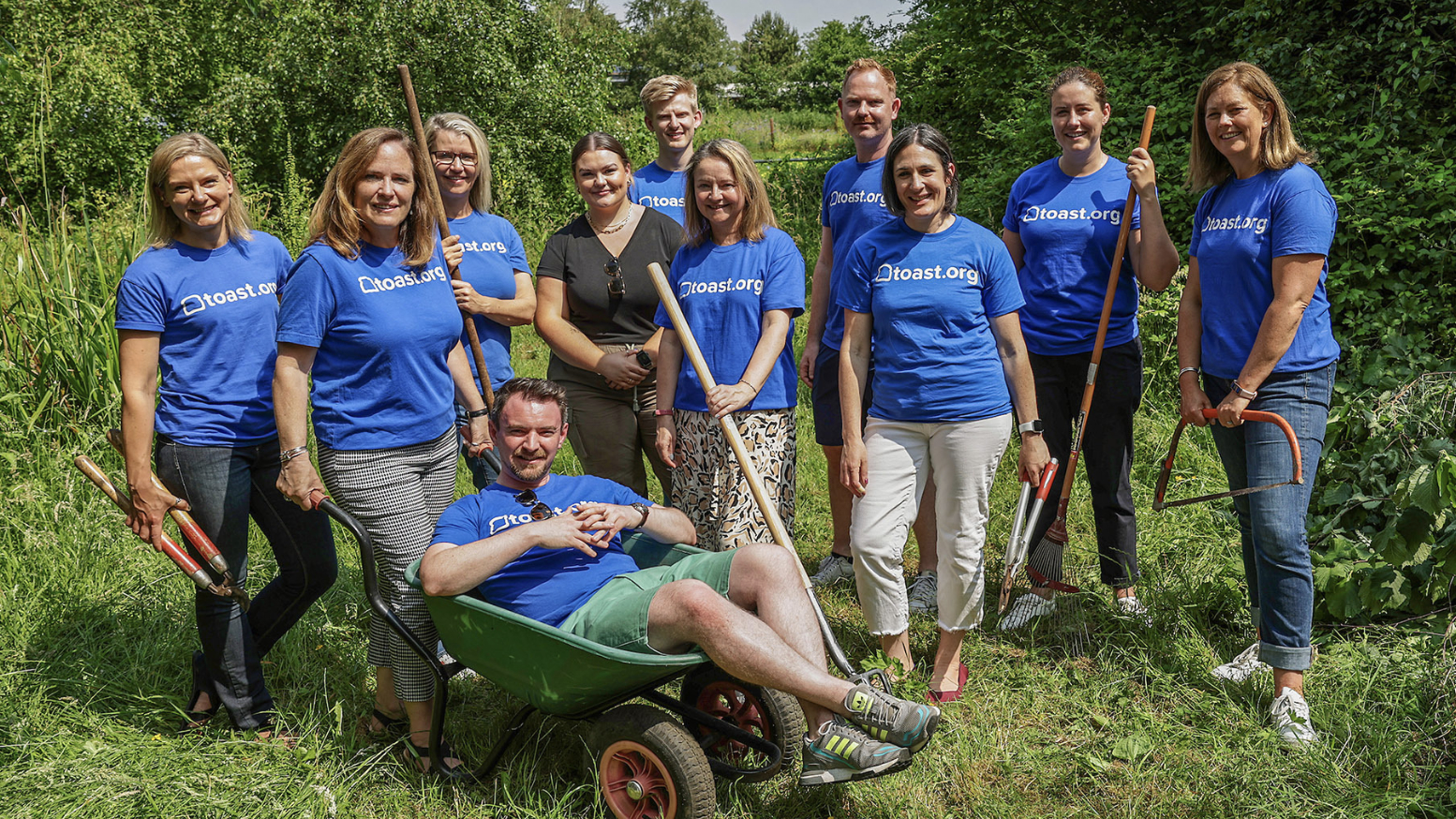 A group of Toasters in Ireland wearing company-branded T-shirts stand with gardening tools in a field, ready for a volunteer event with the company’s philanthropic and social impact branch, Toast.org.  