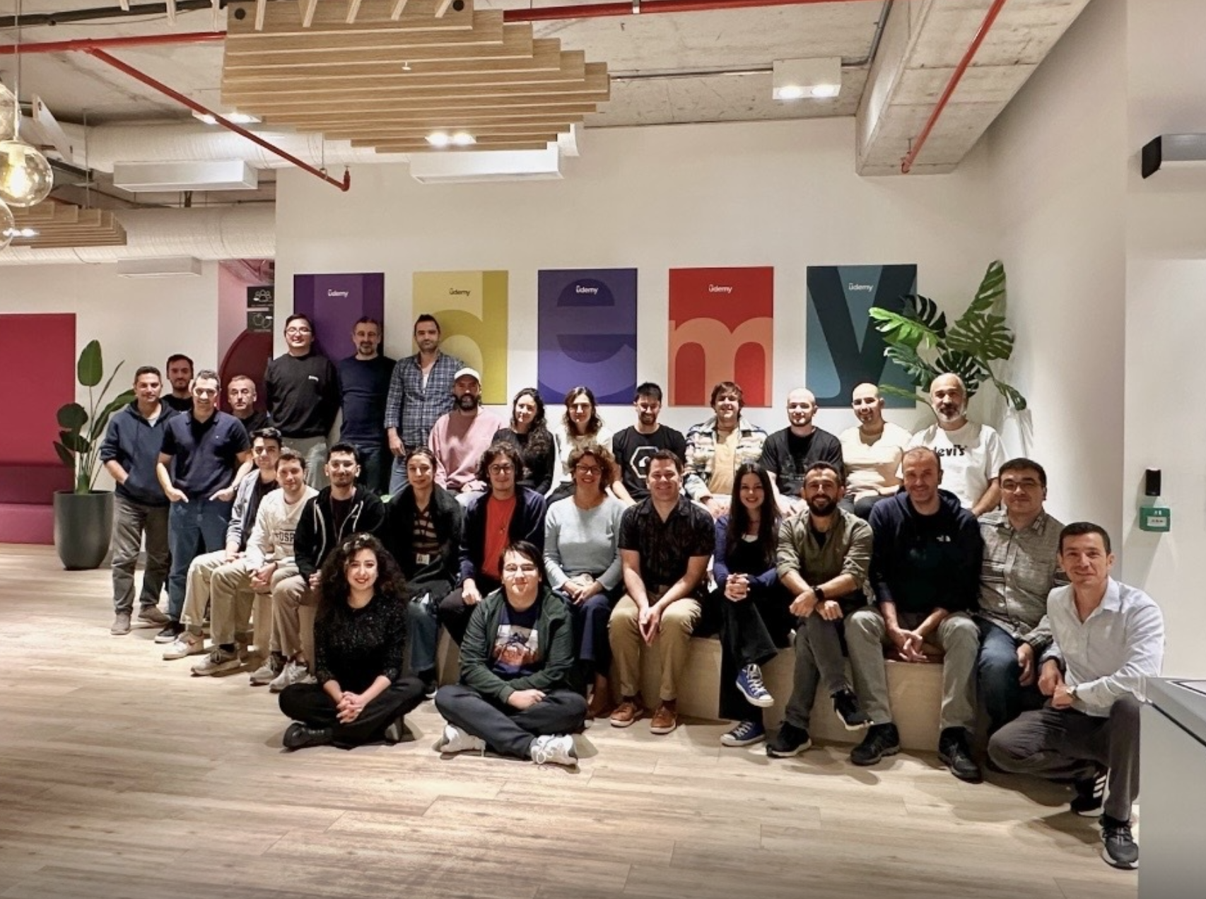 A large group of Udemy employees pose in front of an office wall with a five-panel sign that reads “Udemy.”