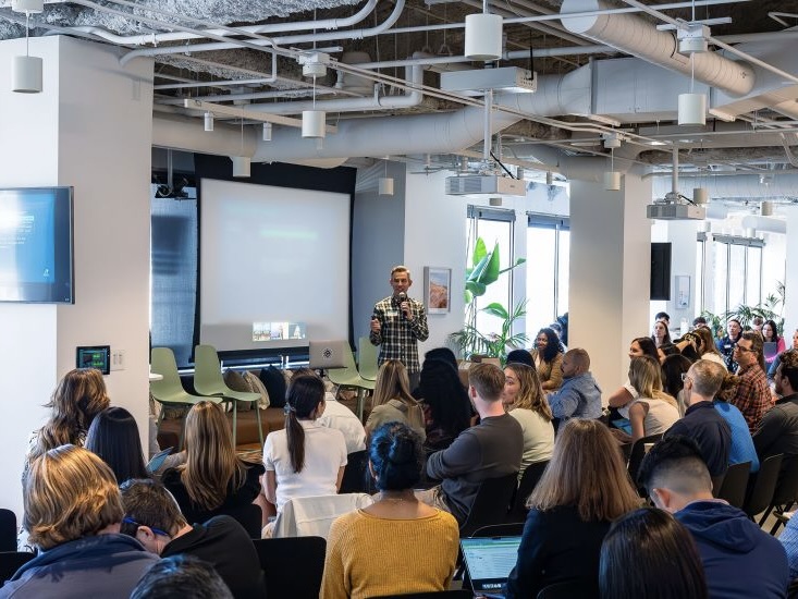 A Grammarly employee speaks into a microphone and presents a slideshow in front of a large seated crowd at a recent meeting.