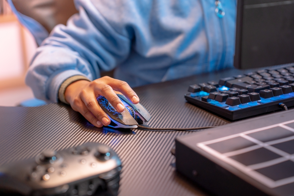 Close range photo of a person’s hand on a light up mouse connected to a gaming PC. The person is wearing a blue zip-up sweatshirt.