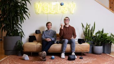 Two Volley teammates sit on a couch, one throwing a small ball in the air and smiling, with the word “Volley” in a neon sign on the wall behind them.