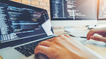 Close-up photo of software developer's hands on laptop keys with two computer screens in background showing code