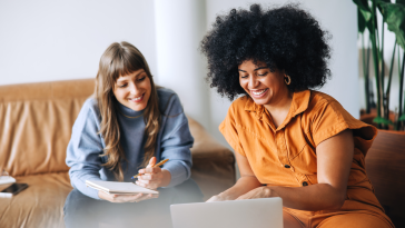 Two young professional women sitting next to each other in a casual work environment looking at a laptop together.