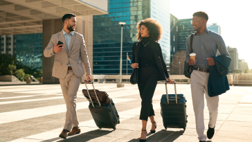 Business team of three walks through an airport parking lot pulling suitcases and drinking coffee